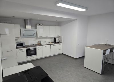 Küche mit weißer Küchenzeile, Backofen, Mikrowelle und Kaffeemaschine, daneben ein Stehtisch mit Barhockern.